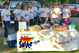 Mora First Citizens employees dressed in white short sleeve tshirts behind a Power Saver table